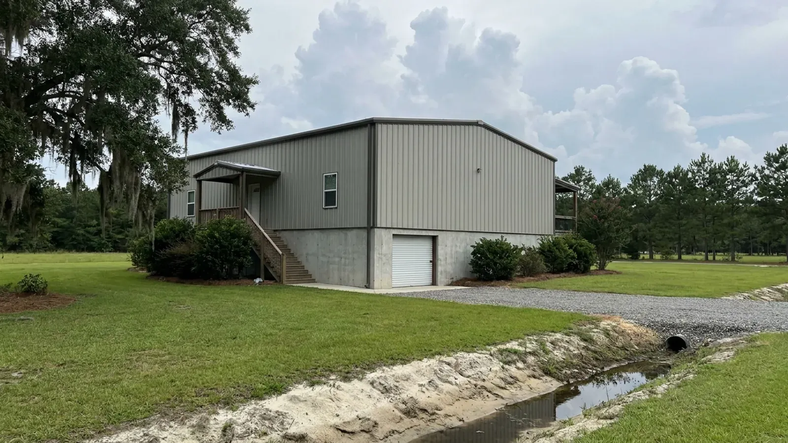 A barndominium raised on a stem-wall foundation with steps up to the entry and enclosed storage space beneath the elevated floor, on flat ground beside a drainage ditch.