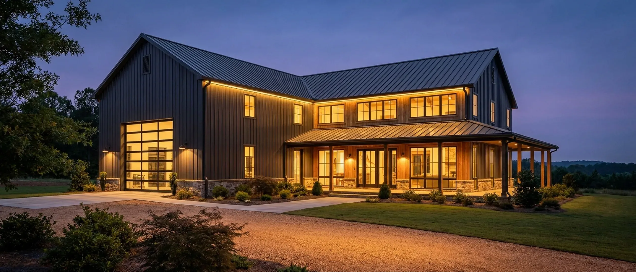 A barndominium at dusk with warm light glowing from every window and exterior lighting along the porch and roofline against a deep blue twilight sky.
